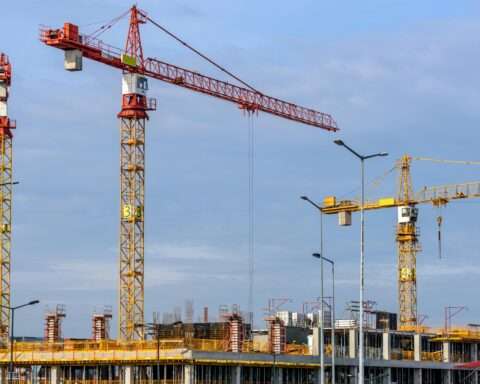 Cranes tower over a construction site with scaffolding before a blue sky.