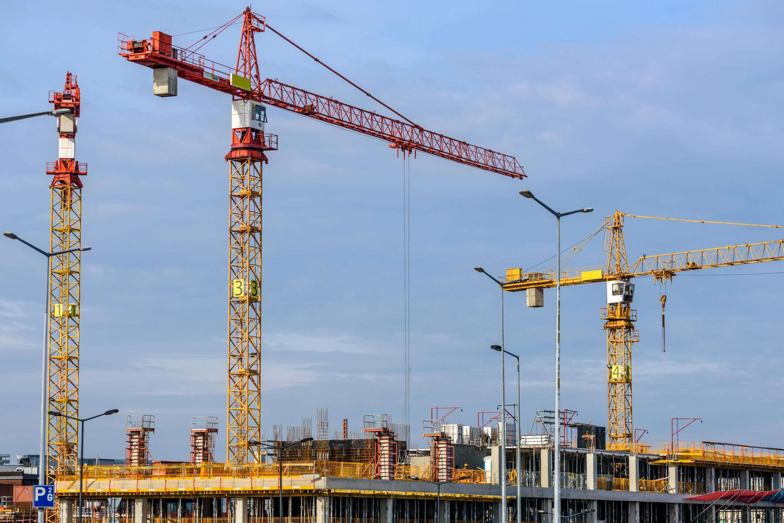 Cranes tower over a construction site with scaffolding before a blue sky.