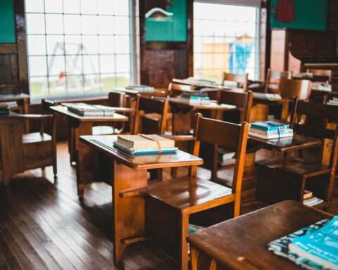 An empty school classroom with wooden desks and books before two windows to the outside world.