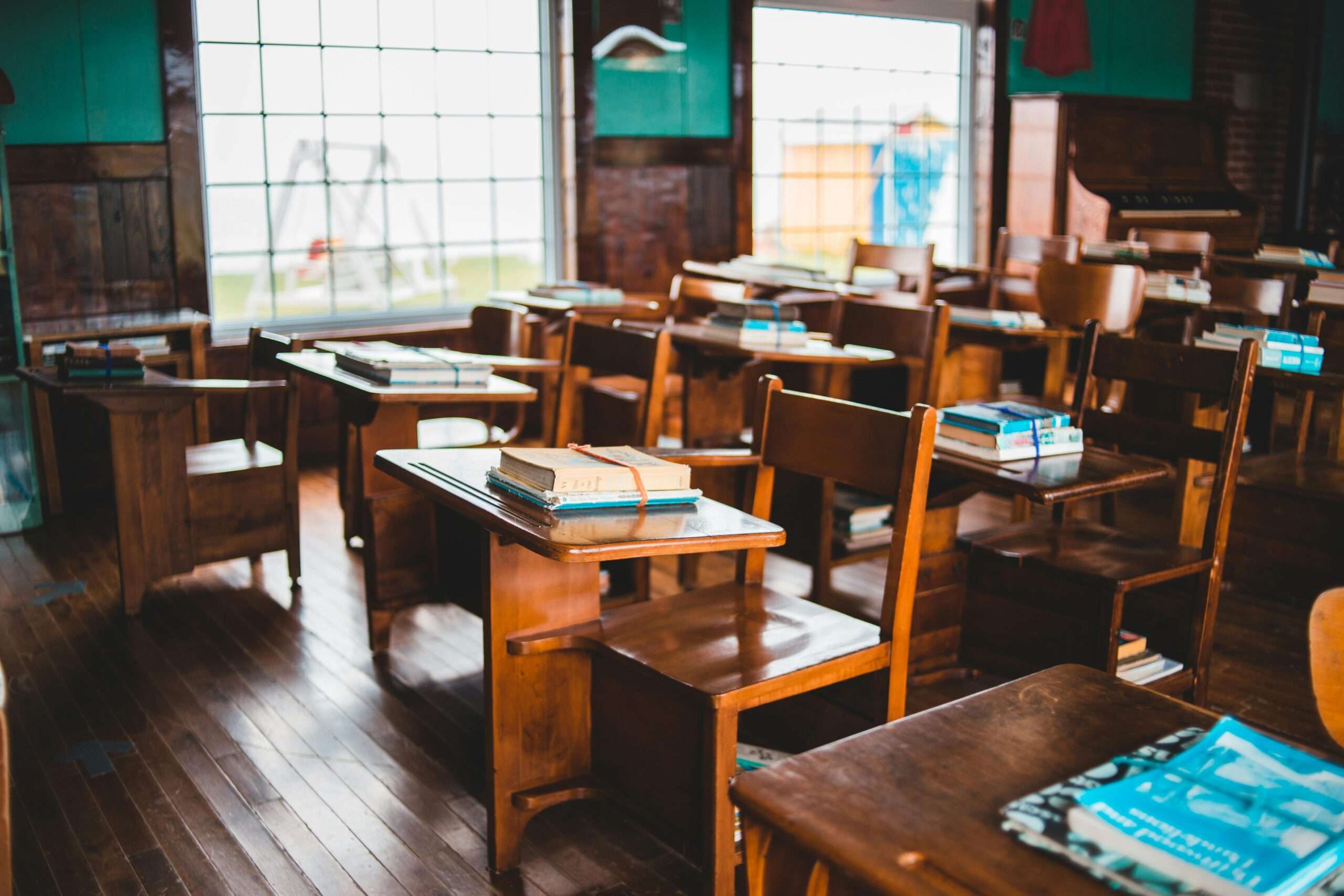 An empty school classroom with wooden desks and books before two windows to the outside world.