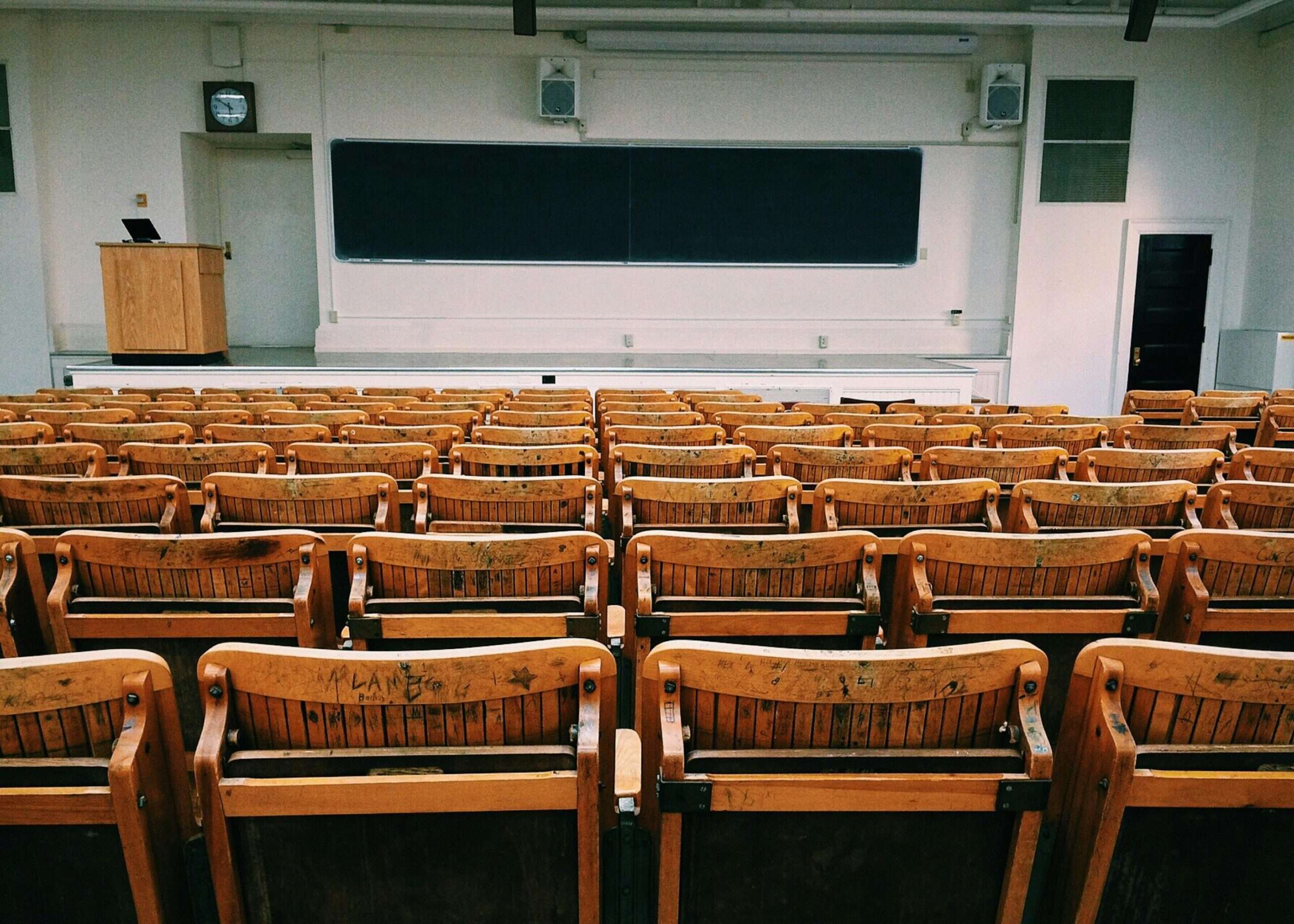 A university classroom with wooden chairs before a black chalkboard.