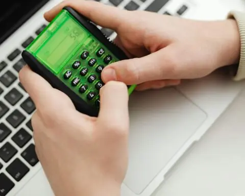A pair of hands types on a calculator before a laptop.
