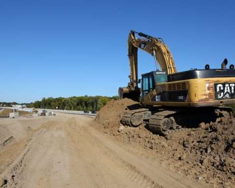 A yellow excavator on a dirt mound alongside a road under construction.