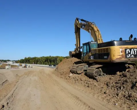 A yellow excavator on a dirt mound alongside a road under construction.