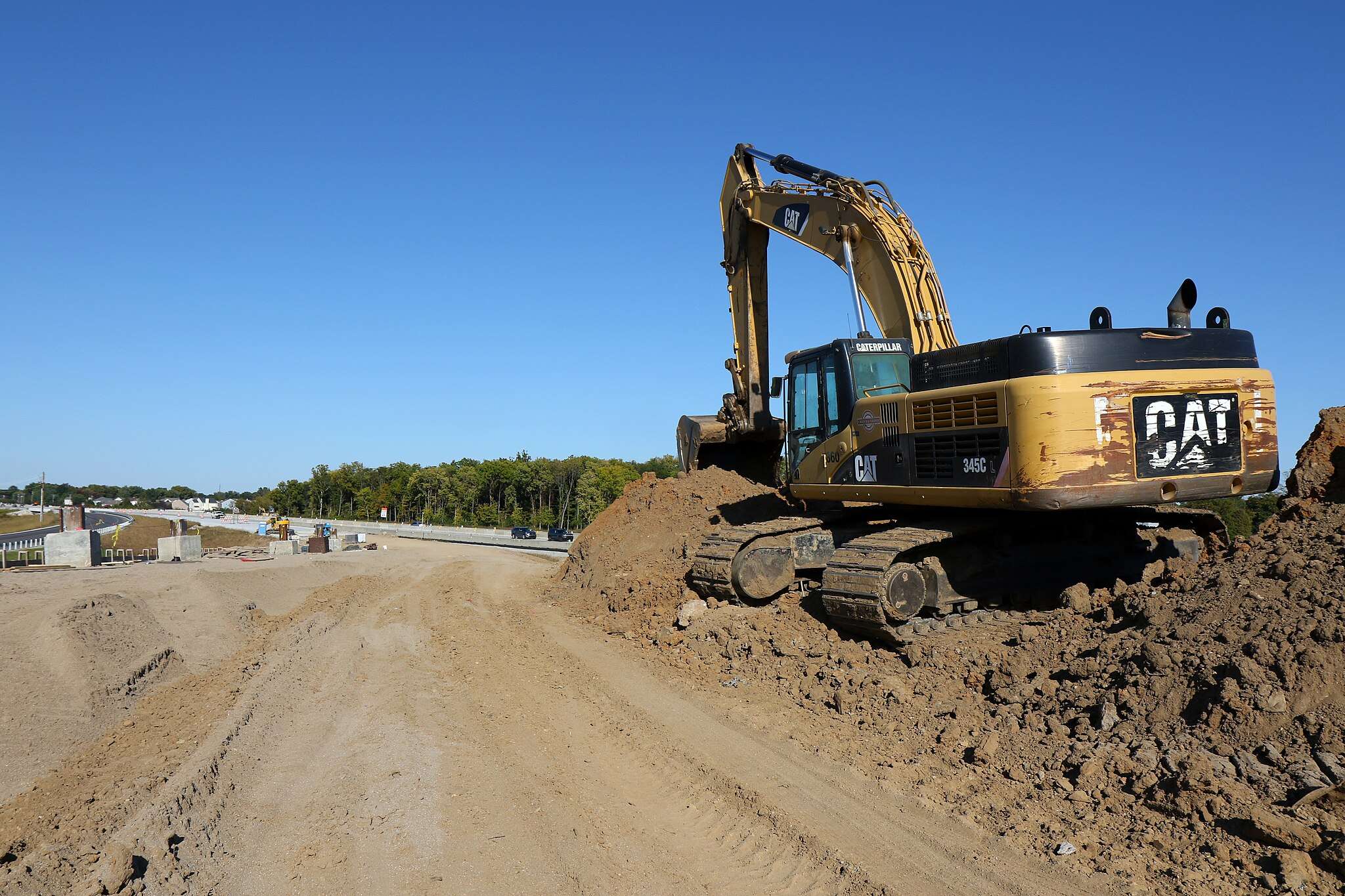 A yellow excavator on a dirt mound alongside a road under construction.