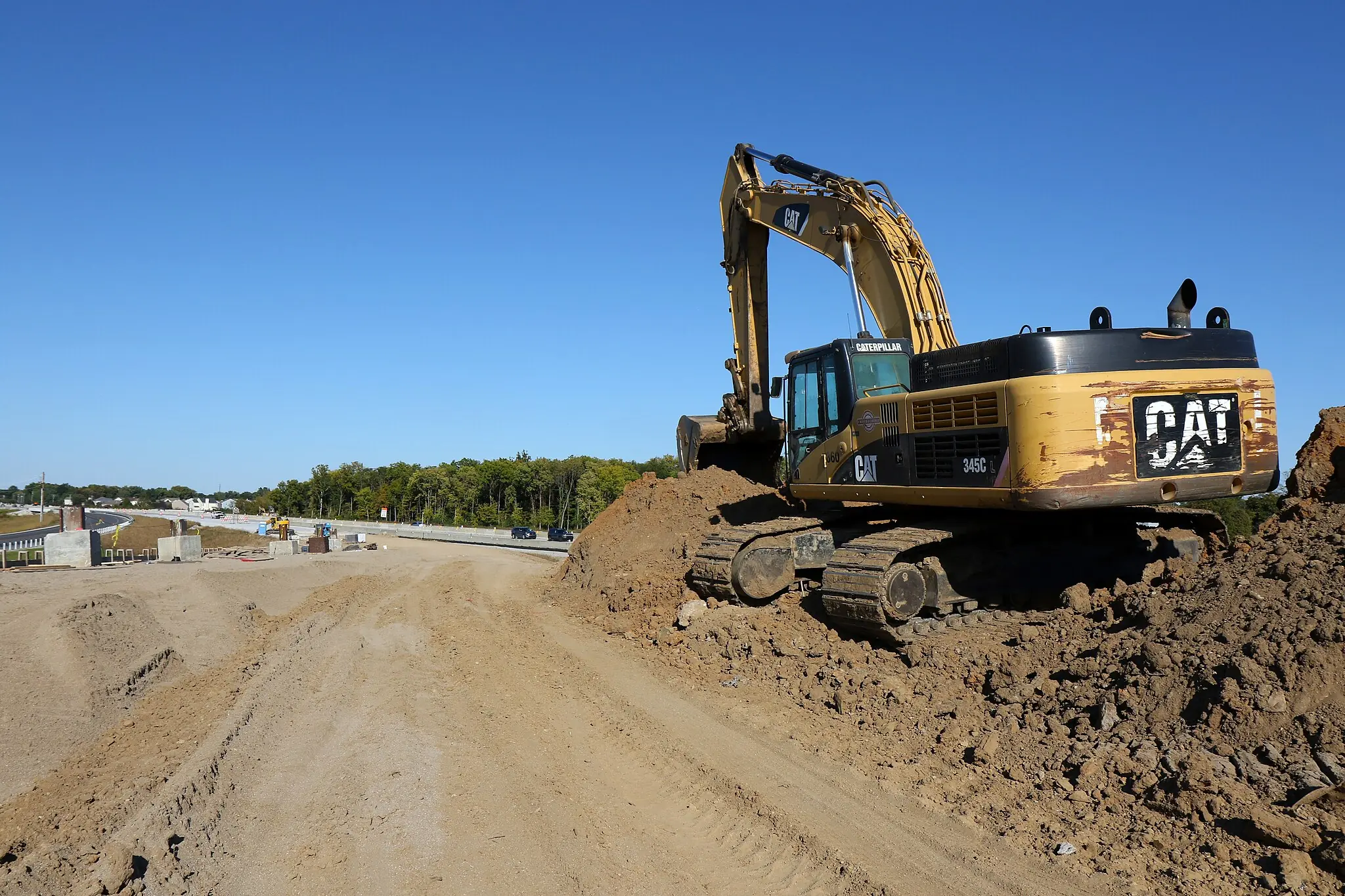 A yellow excavator on a dirt mound alongside a road under construction.