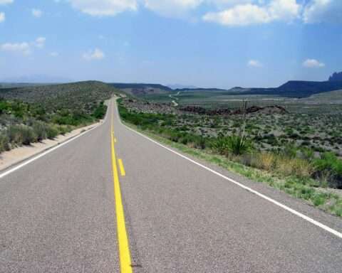 A Texas highway alongside green grass and plains and before a blue sky with clouds.