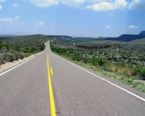 A Texas highway alongside green grass and plains and before a blue sky with clouds.