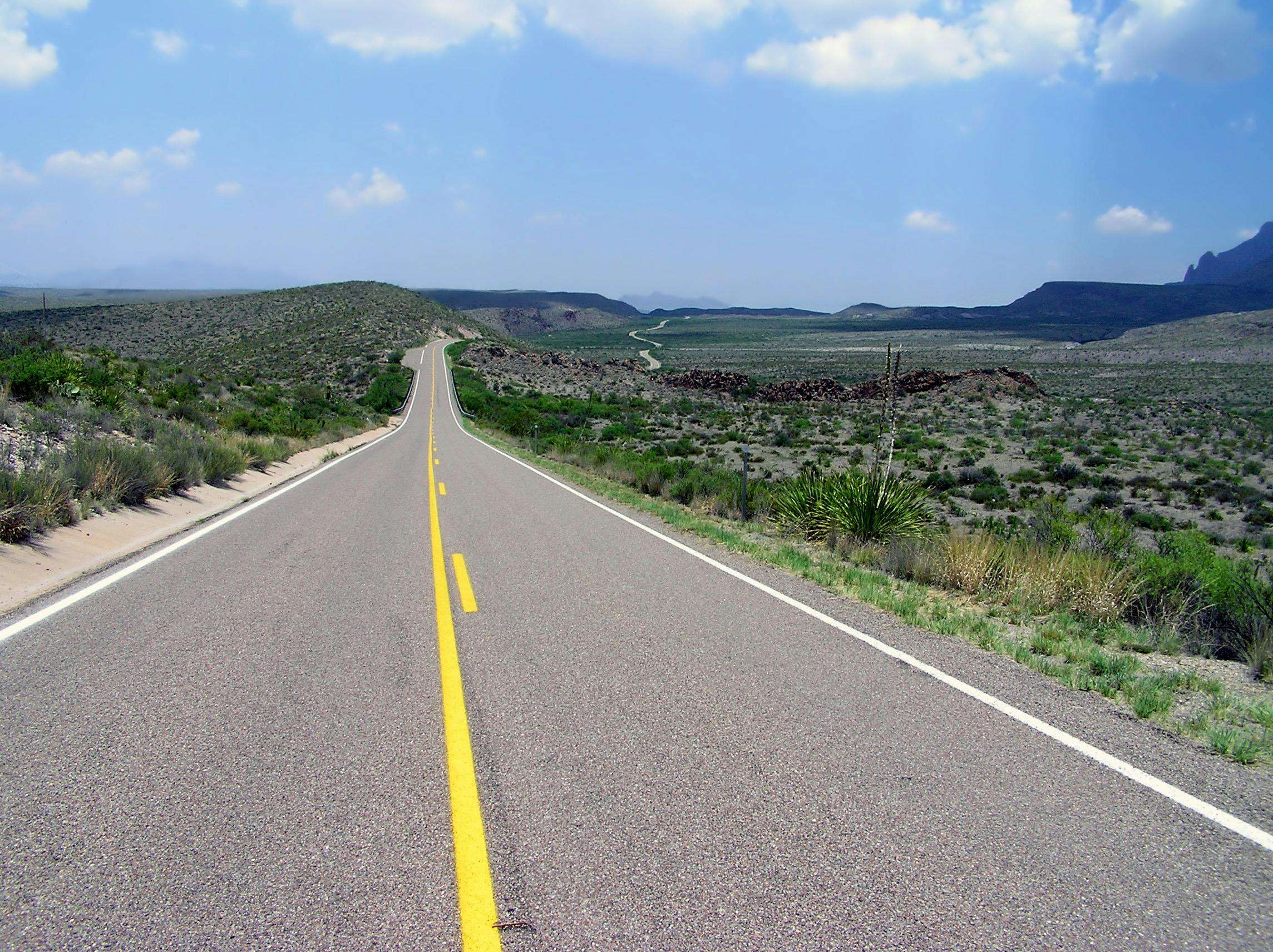 A Texas highway alongside green grass and plains and before a blue sky with clouds.