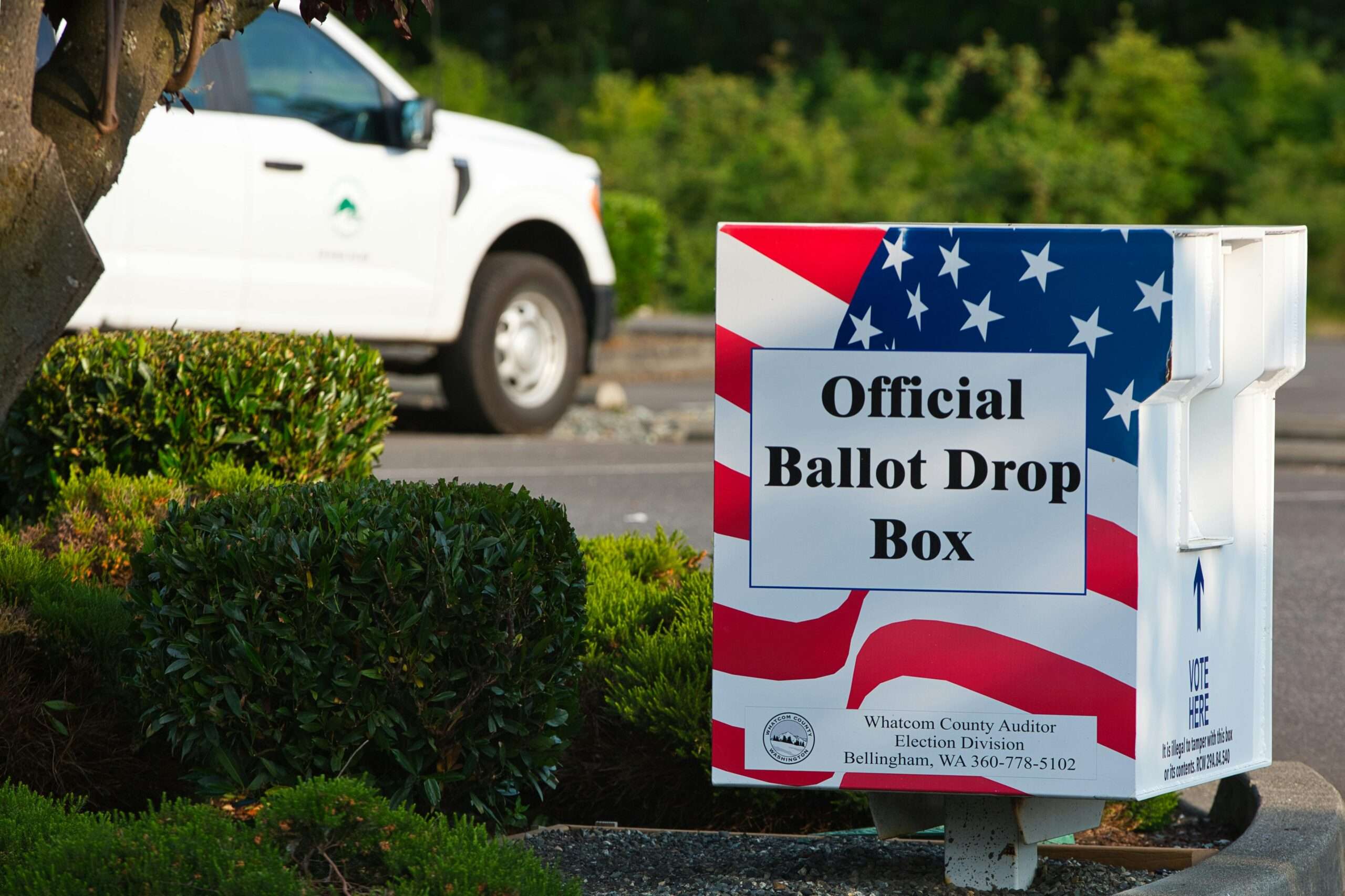 A red, white and blue ballot box before green grass and a parked car.