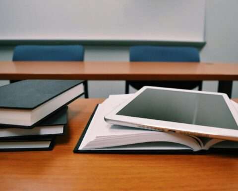 A tablet and school books on a desk in an empty classroom with a whiteboard.