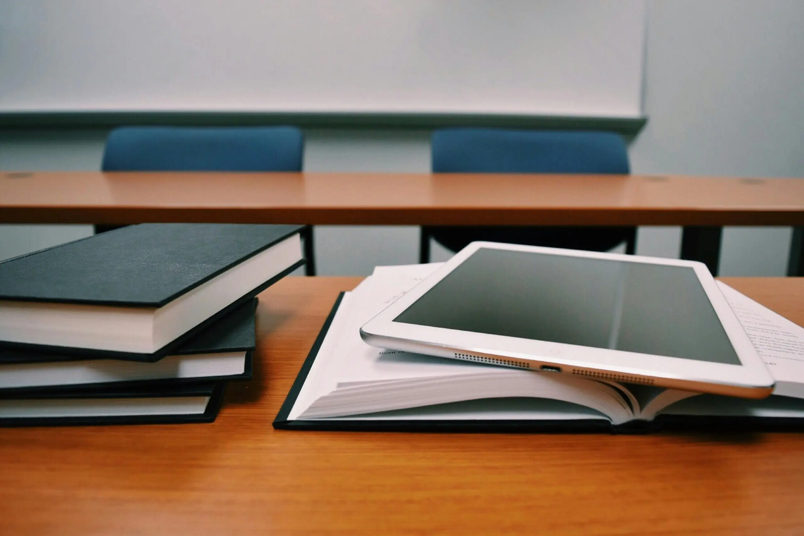 A tablet and school books on a desk in an empty classroom with a whiteboard.