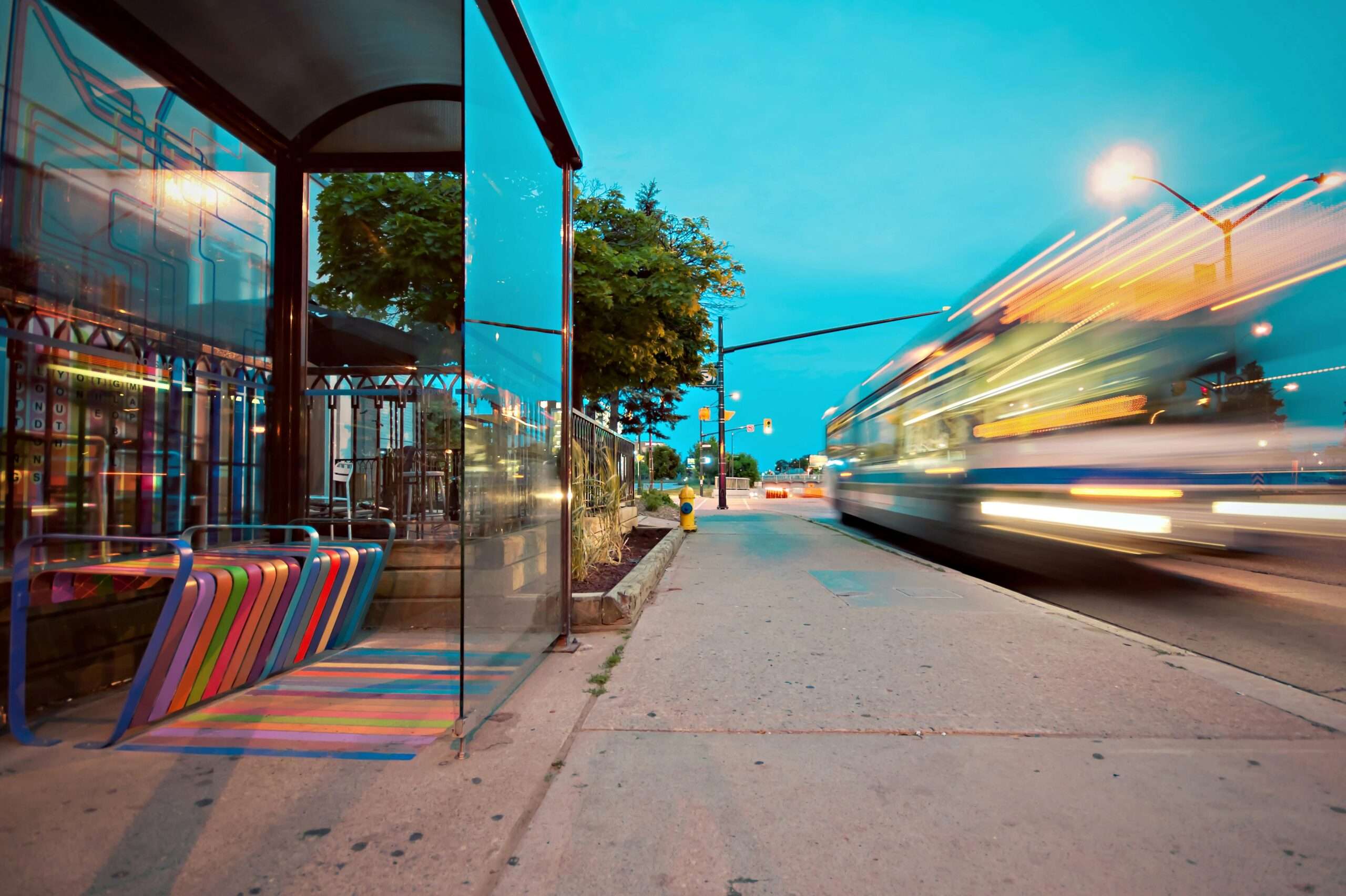 A bus zooms past a bus station with a multi-colored bench.