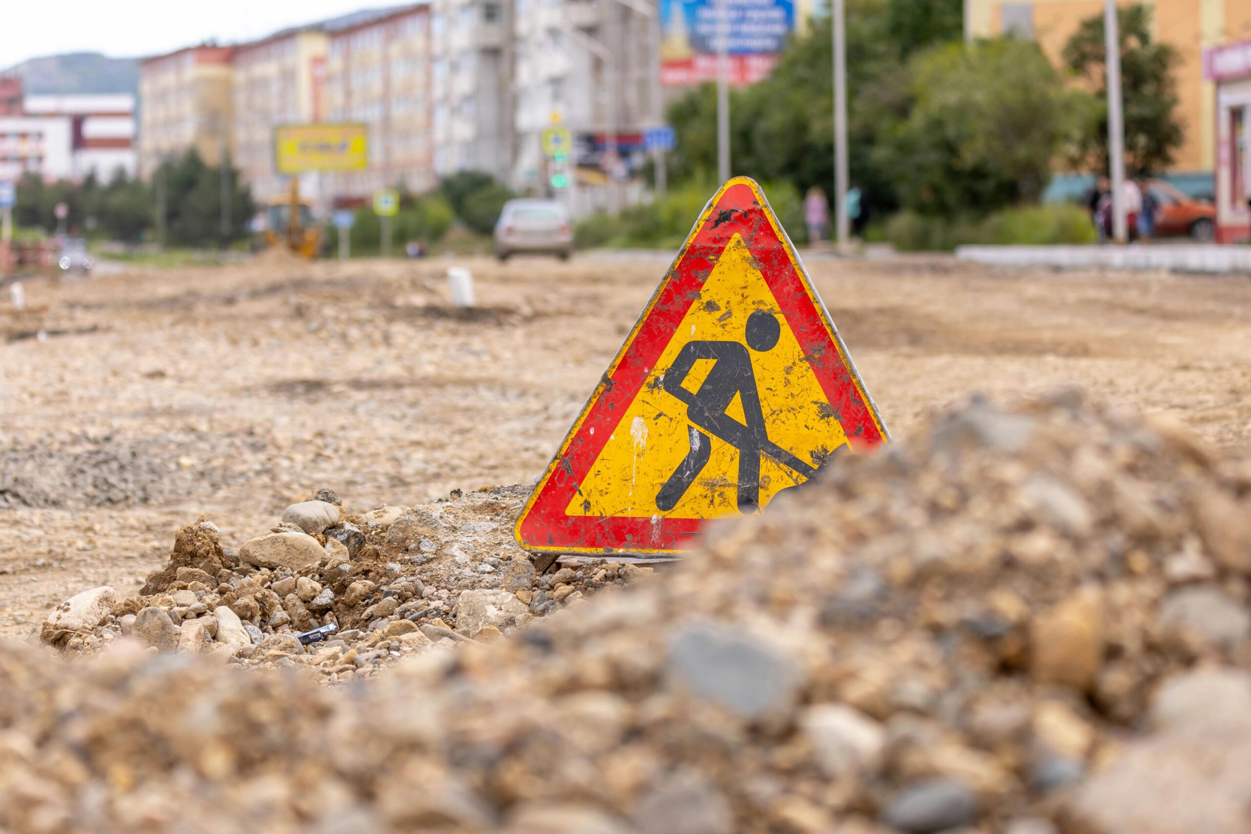 A road work sign on a gravel and dirt patch in an urban area.