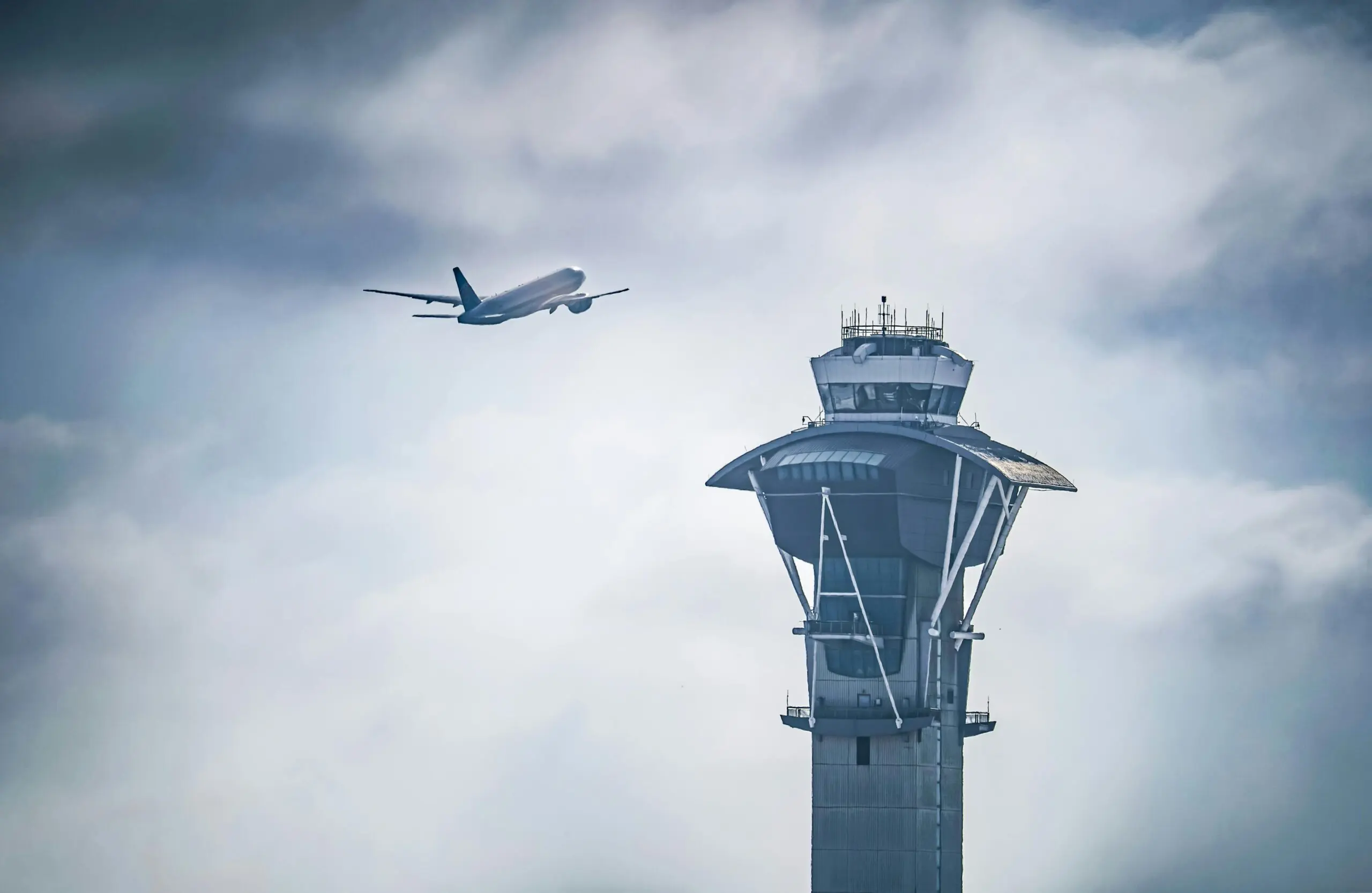An air traffic control tower before a flying plane in the distance and a blue sky.