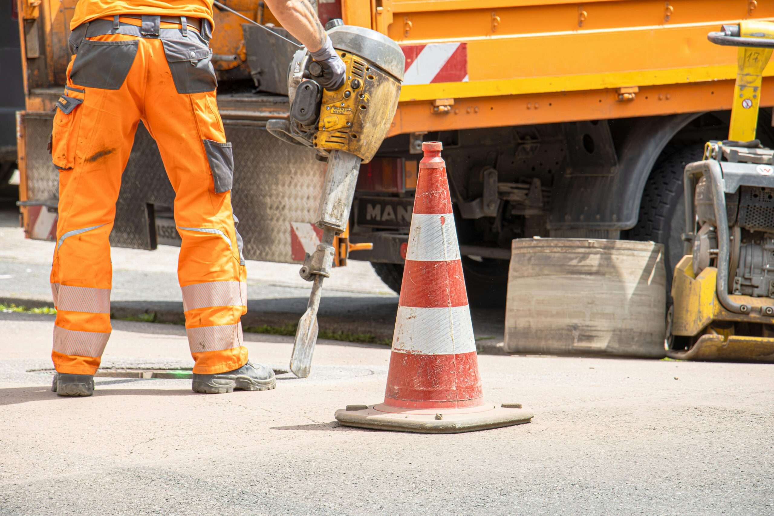 The bottom half of a construction worker in high-vis gear nearby an orange traffic cone and vehicle.