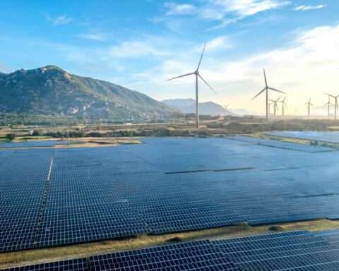 A lot of solar panels before wind turbines and a grass hill on a sunny day.