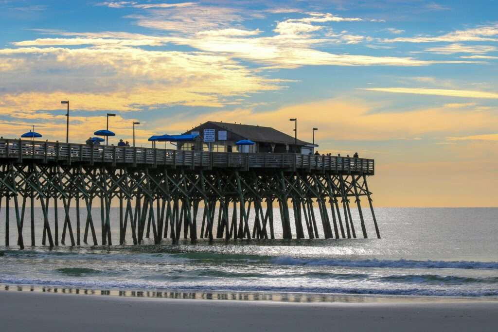 A storm damaged beach before a pier with a structure above rolling waves.