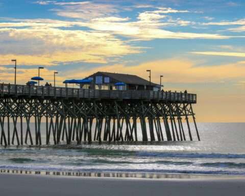 A storm damaged beach before a pier with a structure above rolling waves.