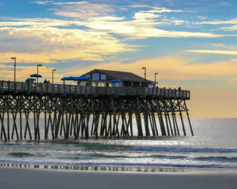 A storm damaged beach before a pier with a structure above rolling waves.