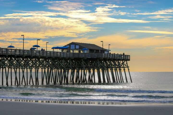 A storm damaged beach before a pier with a structure above rolling waves.