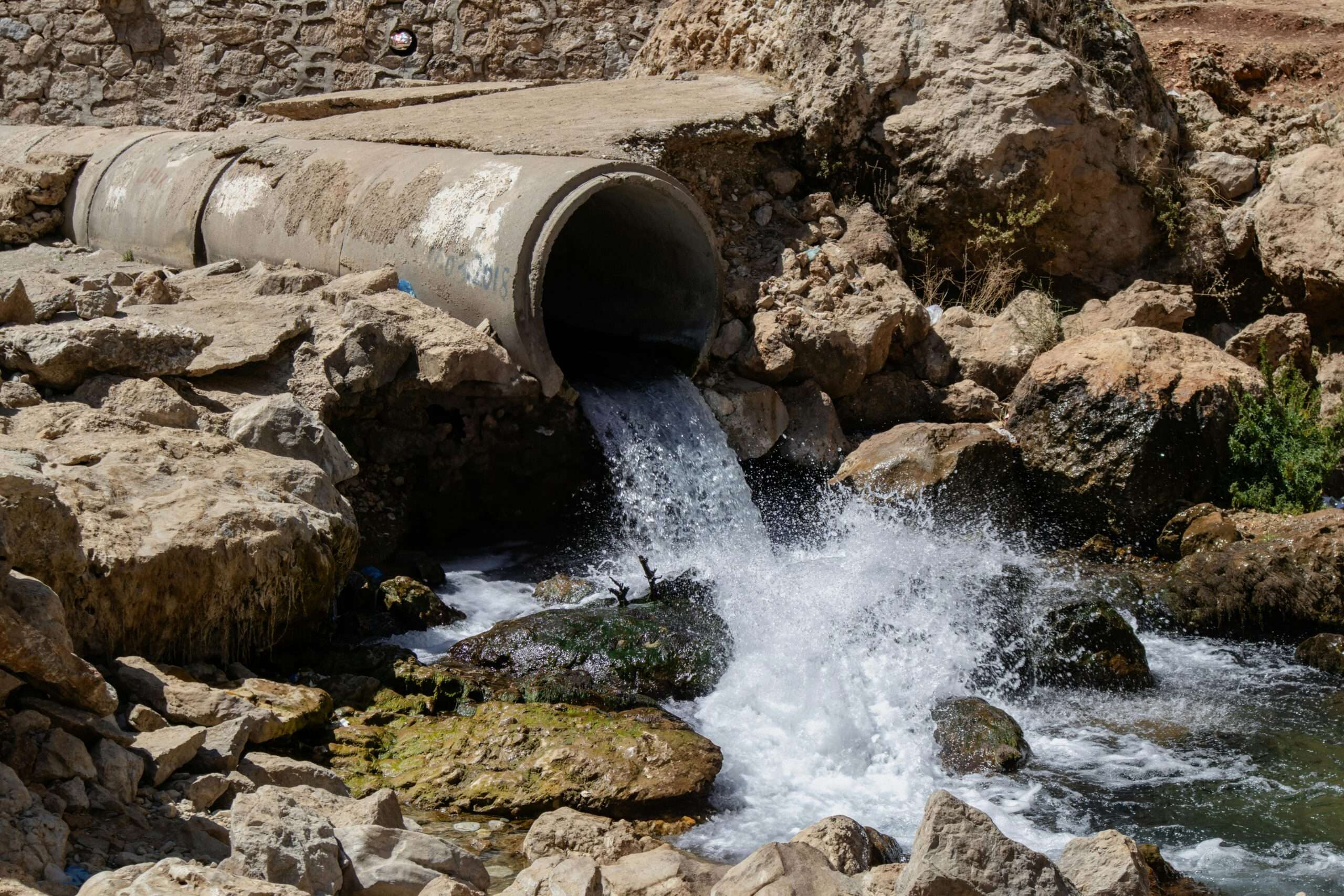 Water pours from a wastewater pipe into rocks before a scenic park background.