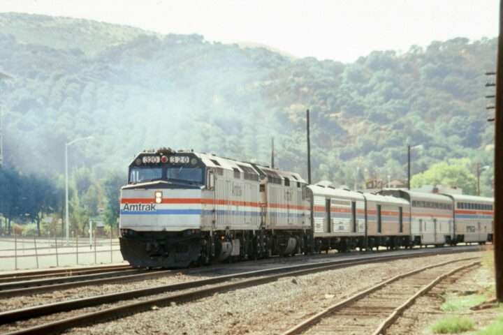 An Amtrak train on tracks before a green hill with trees and bushes.