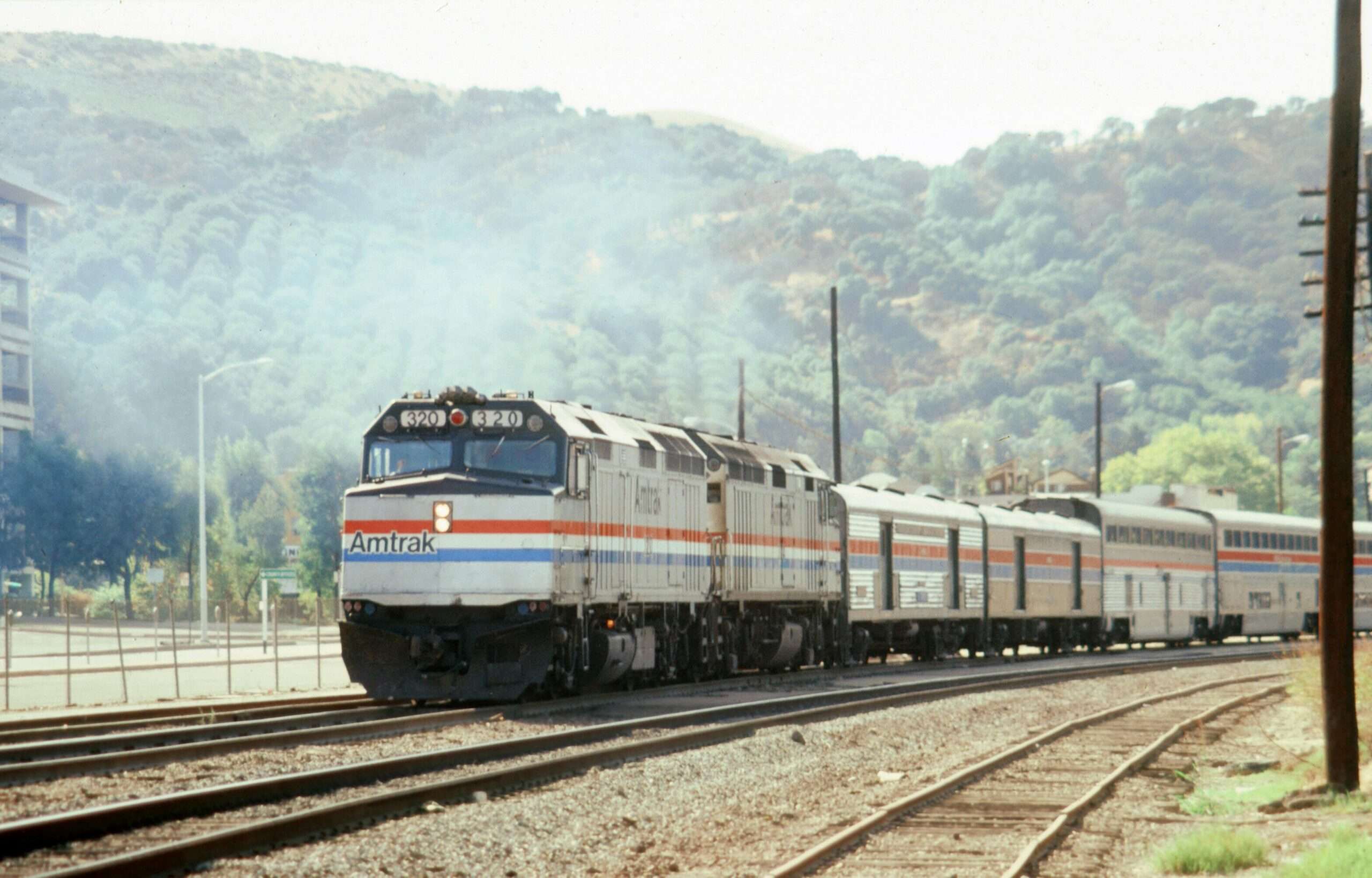An Amtrak train on tracks before a green hill with trees and bushes.
