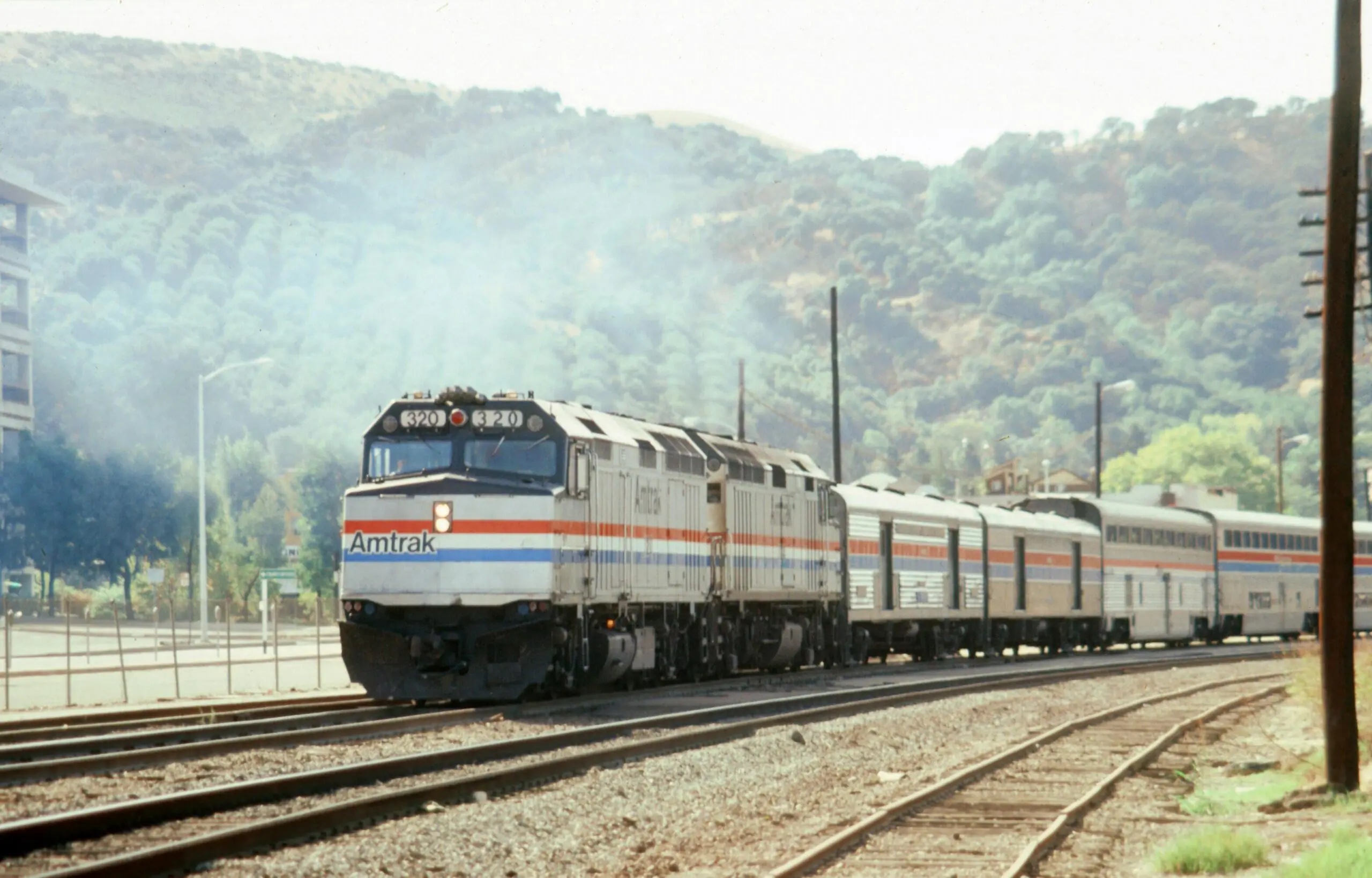An Amtrak train on tracks before a green hill with trees and bushes.